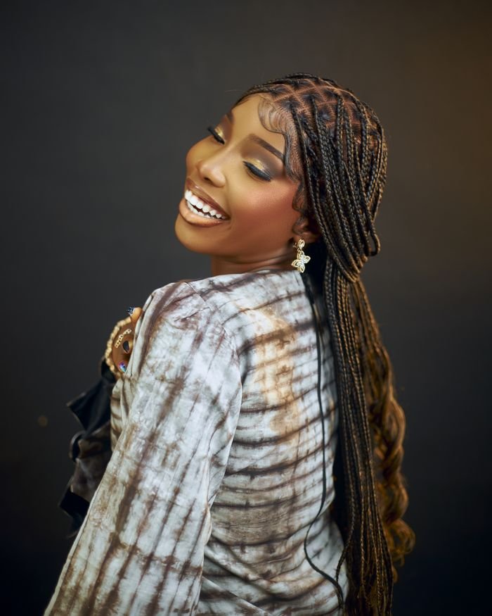 A joyful woman with braided hair smiling happily, wearing tie-dye clothing in a studio setting.