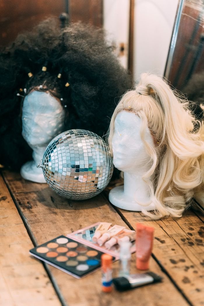 Shimmering silver disco ball between dark and fair hair of wigs composed with different cosmetics on wooden floor with mirror